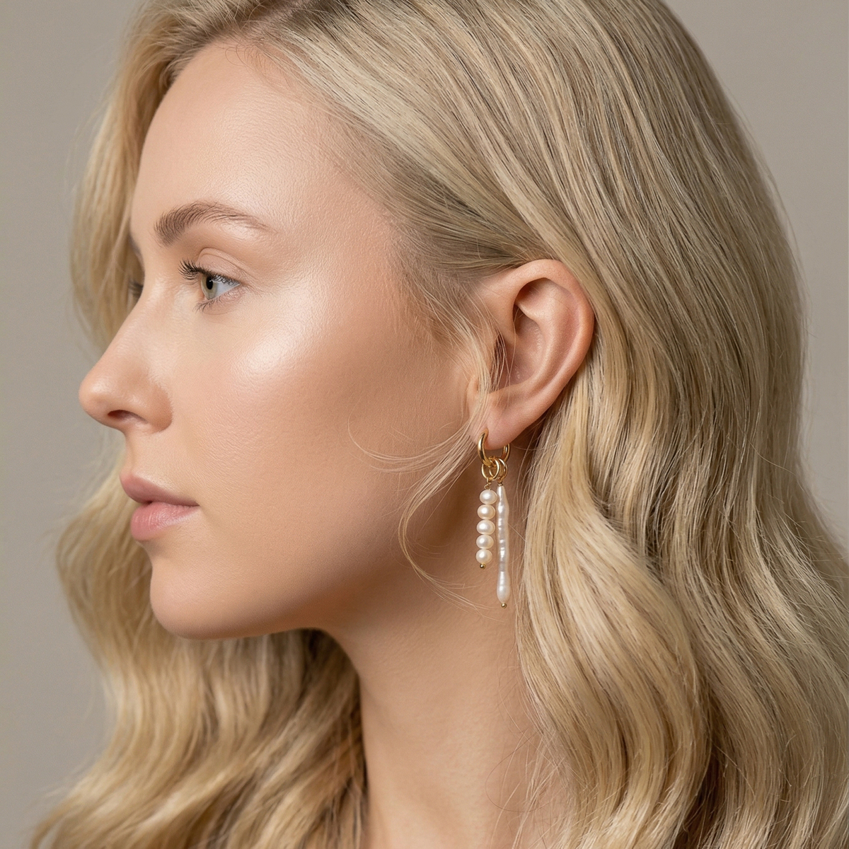 Close-up of a woman wearing an 18k gold plated huggie hoop earring adorned with a long pink baroque stick pearl and a matching five-pearl cluster charm.