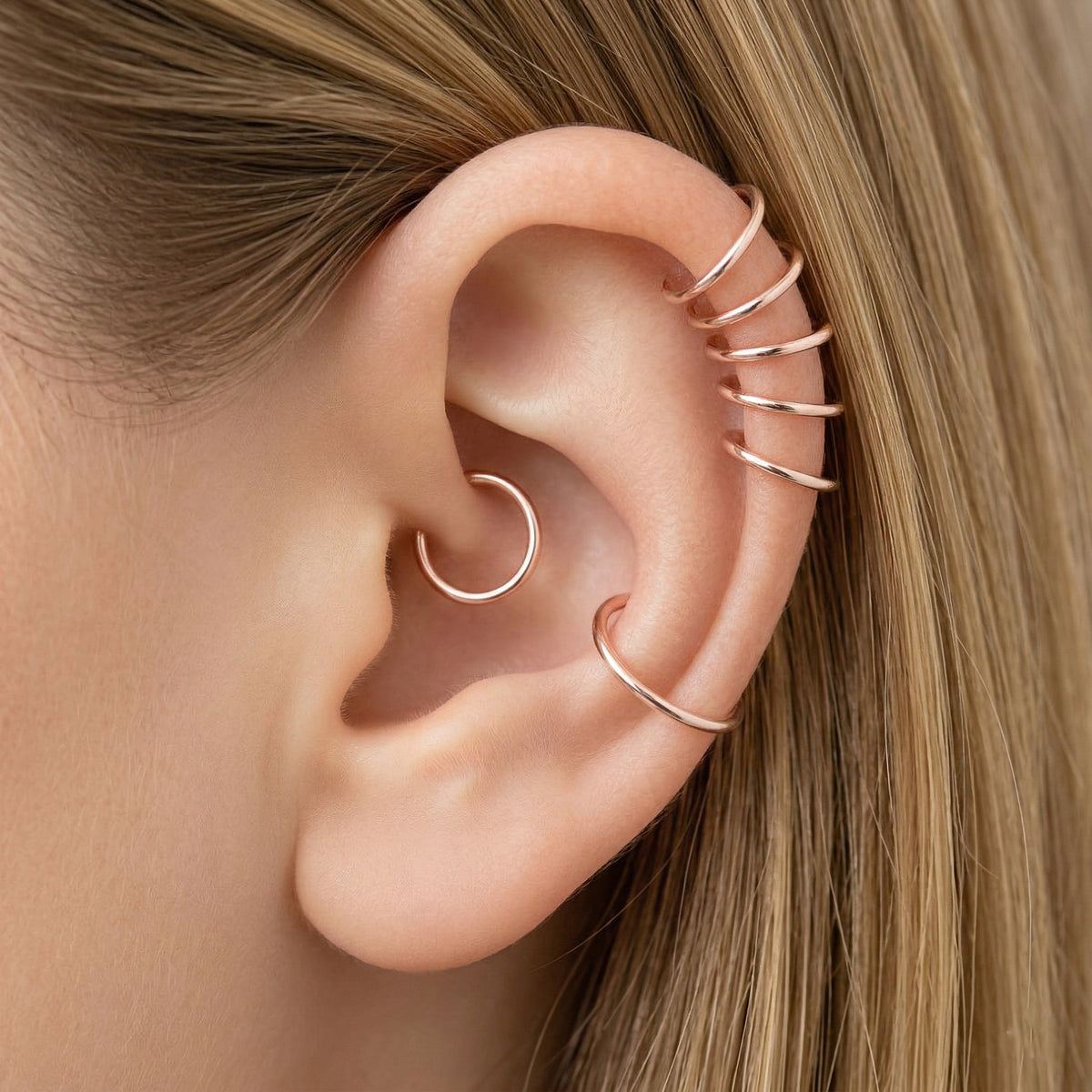 Close-up of a human ear featuring multiple smooth rose gold seamless hoop earrings in various sizes, including helix, tragus, and lobe piercings.