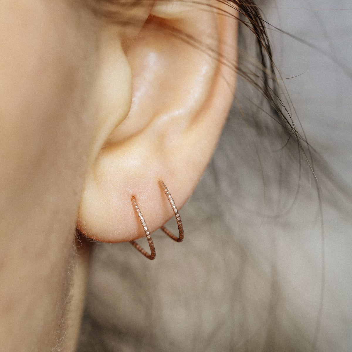 Close-up of a woman's ear wearing tiny rose gold double hoop illusion earrings with a diamond-cut texture, designed to create the appearance of two piercings with a single hole.