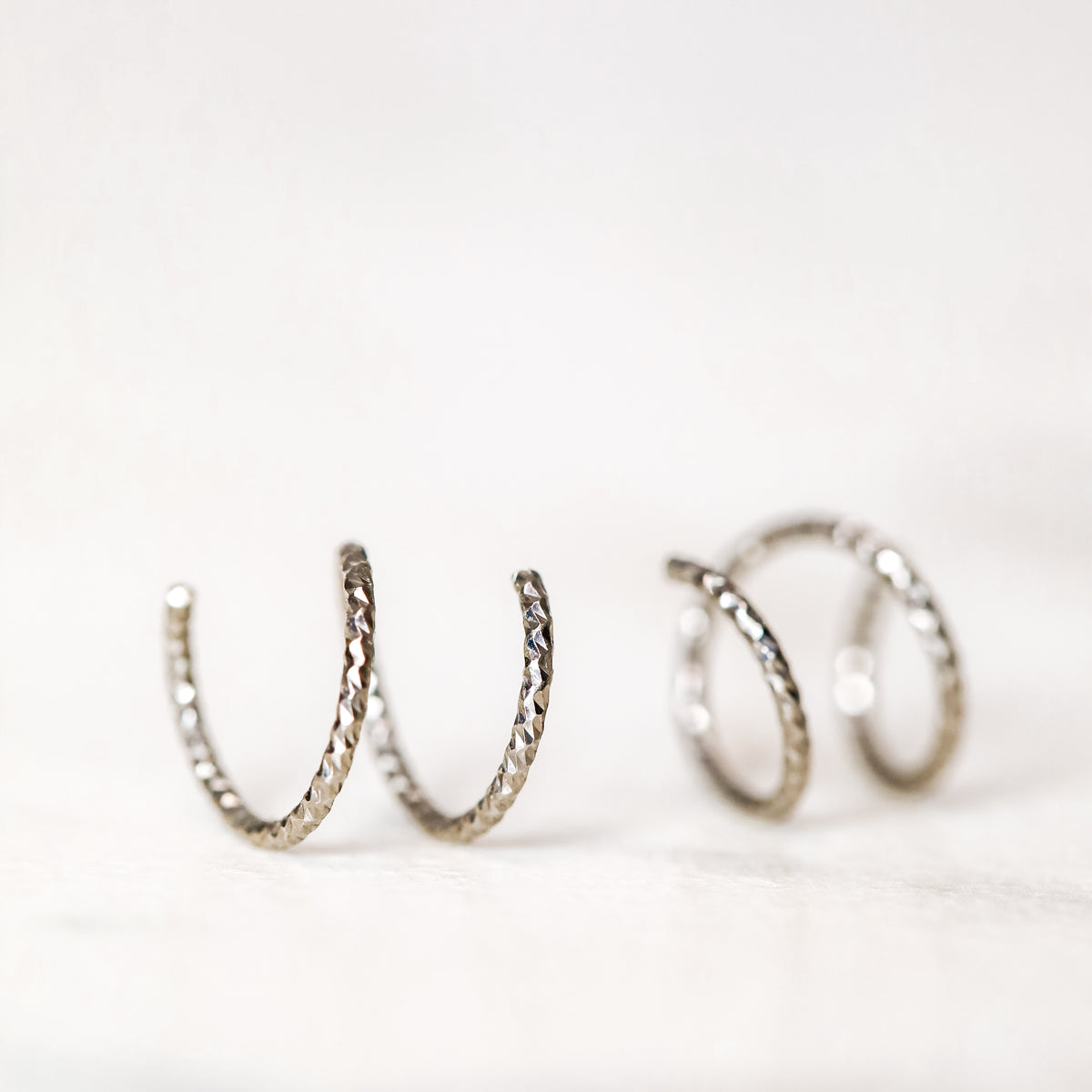 Minimal black double hoop spiral earrings worn on ear, tiny ruthenium sterling silver illusion hoops creating double piercing look, diamond cut texture close-up