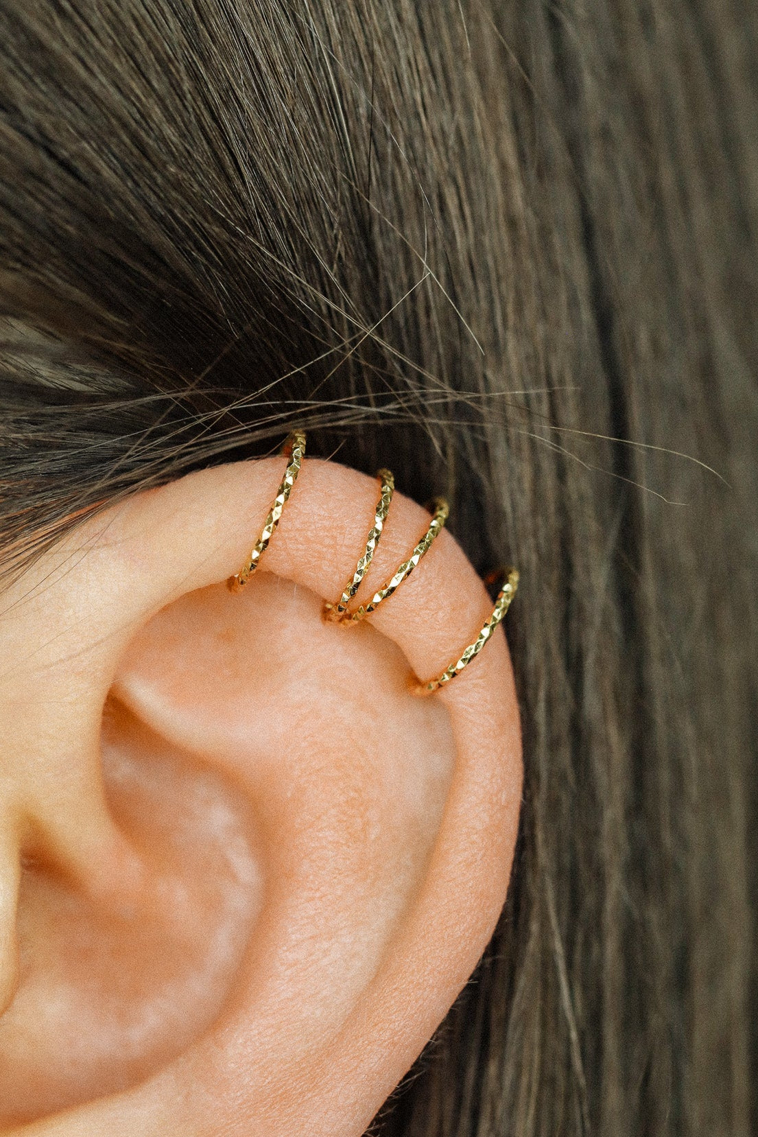 Close-up of a person's ear wearing three textured gold diamond-cut ear cuffs on the helix, showing the adjustable fit and shimmering gold finish.