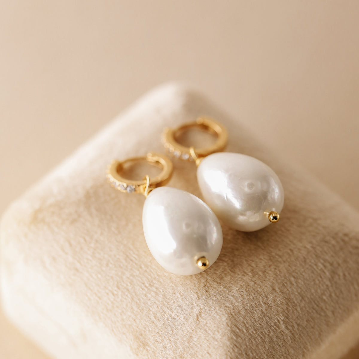 Pair of 18k gold-plated huggie hoop earrings with premium teardrop pearl drops and cubic zirconia crystals, displayed on a beige velvet jewelry box.