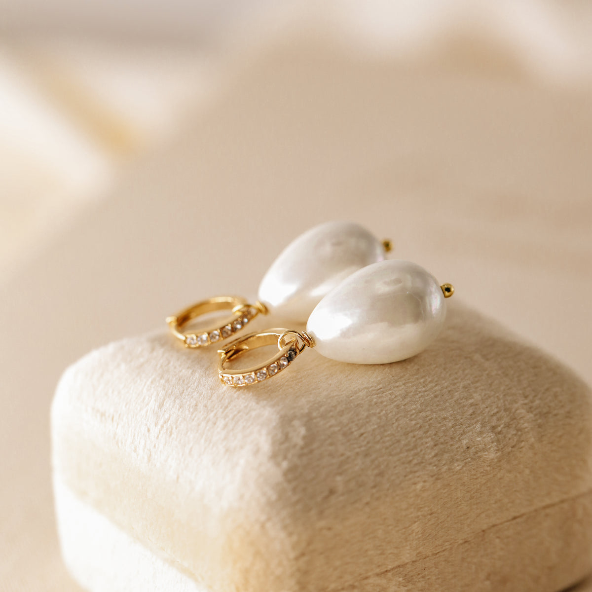Pair of 18k gold-plated huggie hoop earrings with premium teardrop pearl drops and cubic zirconia crystals, displayed on a beige velvet jewelry box.