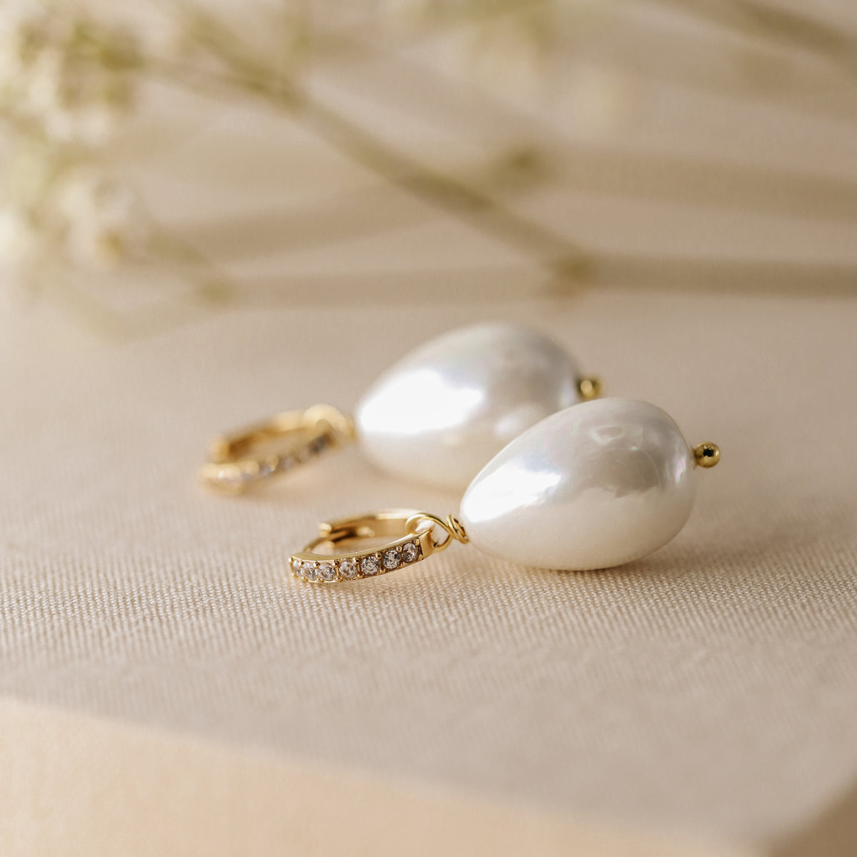 Close-up shot of two handmade teardrop pearl earrings with gold micro-pave hoops on a textured linen surface with soft floral background.