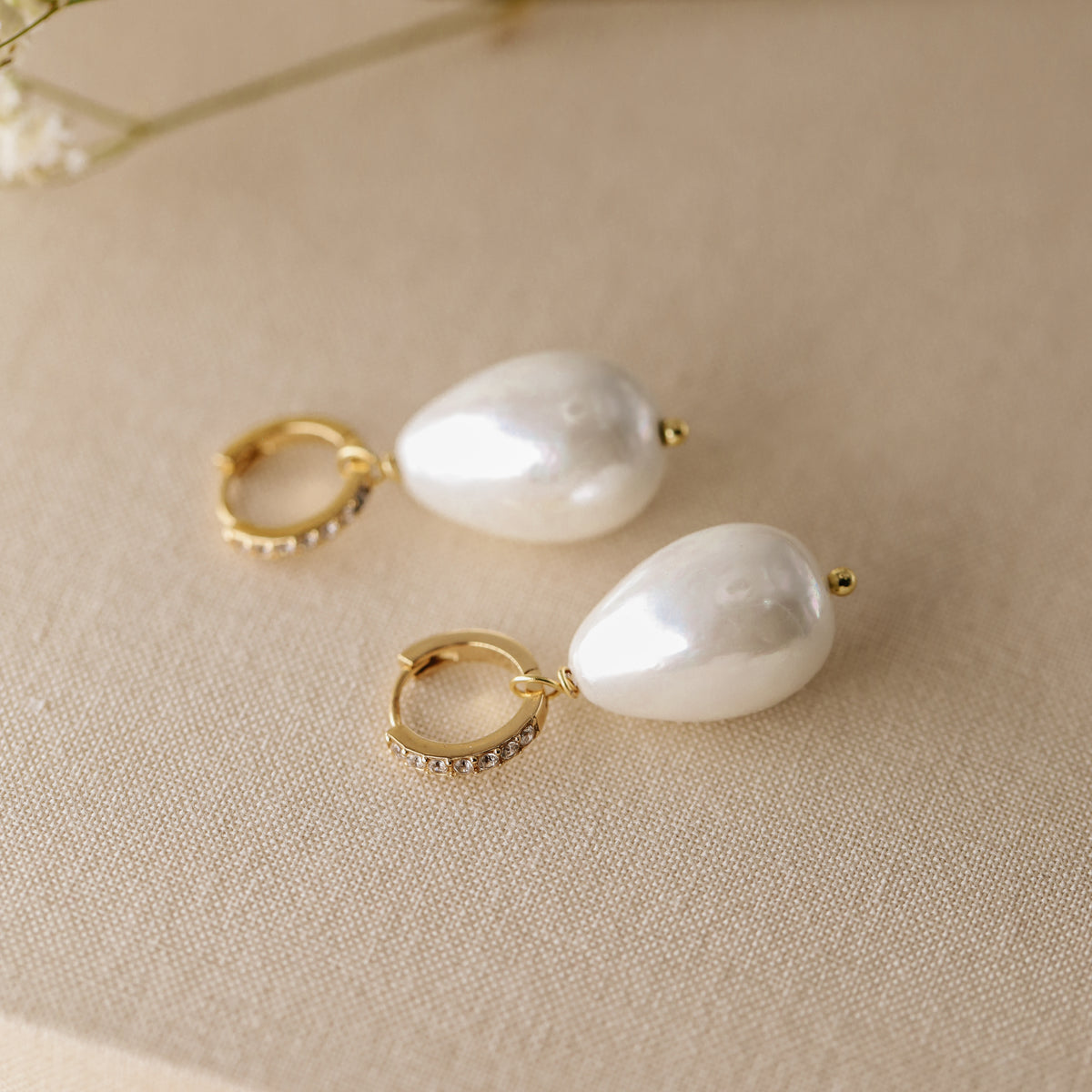 Close-up shot of two handmade teardrop pearl earrings with gold micro-pave hoops on a textured linen surface with soft floral background.