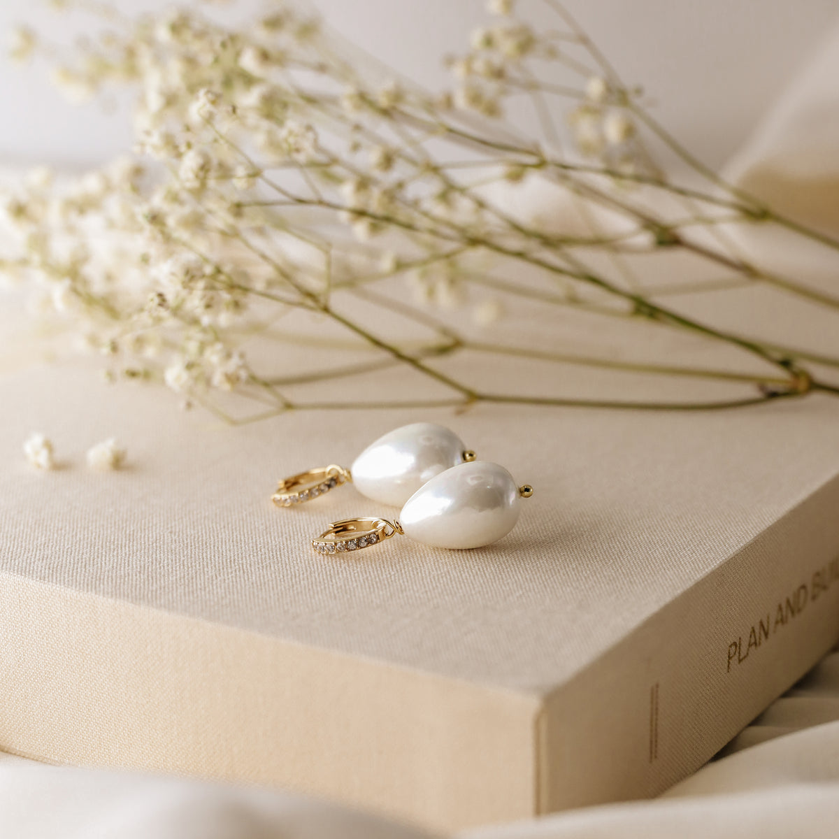 Close-up shot of two handmade teardrop pearl earrings with gold micro-pave hoops on a textured linen surface with soft floral background.