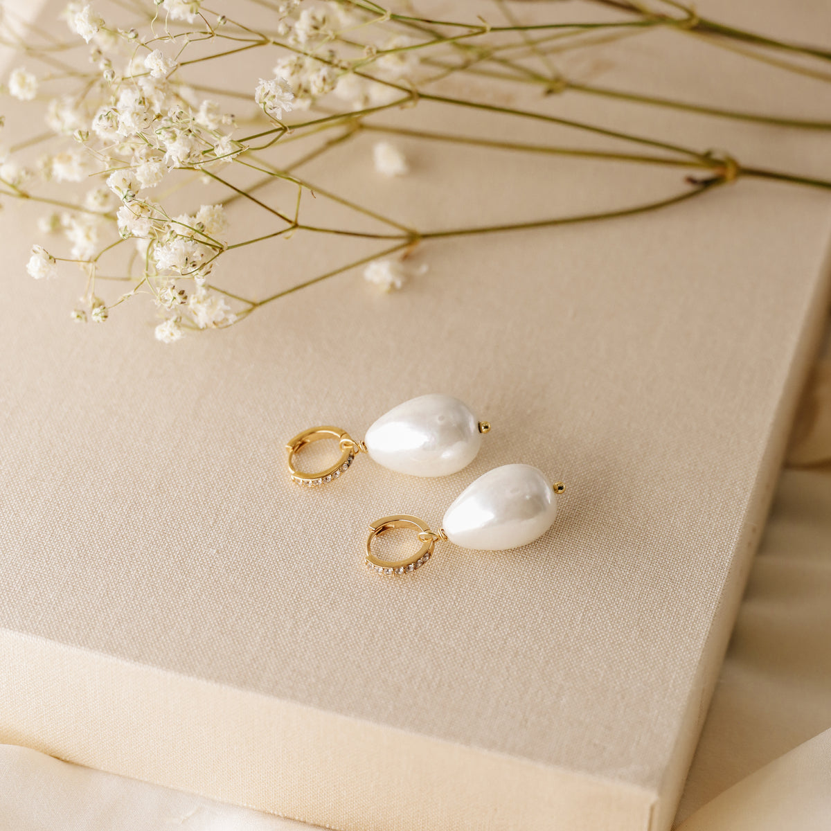 Close-up shot of two handmade teardrop pearl earrings with gold micro-pave hoops on a textured linen surface with soft floral background.