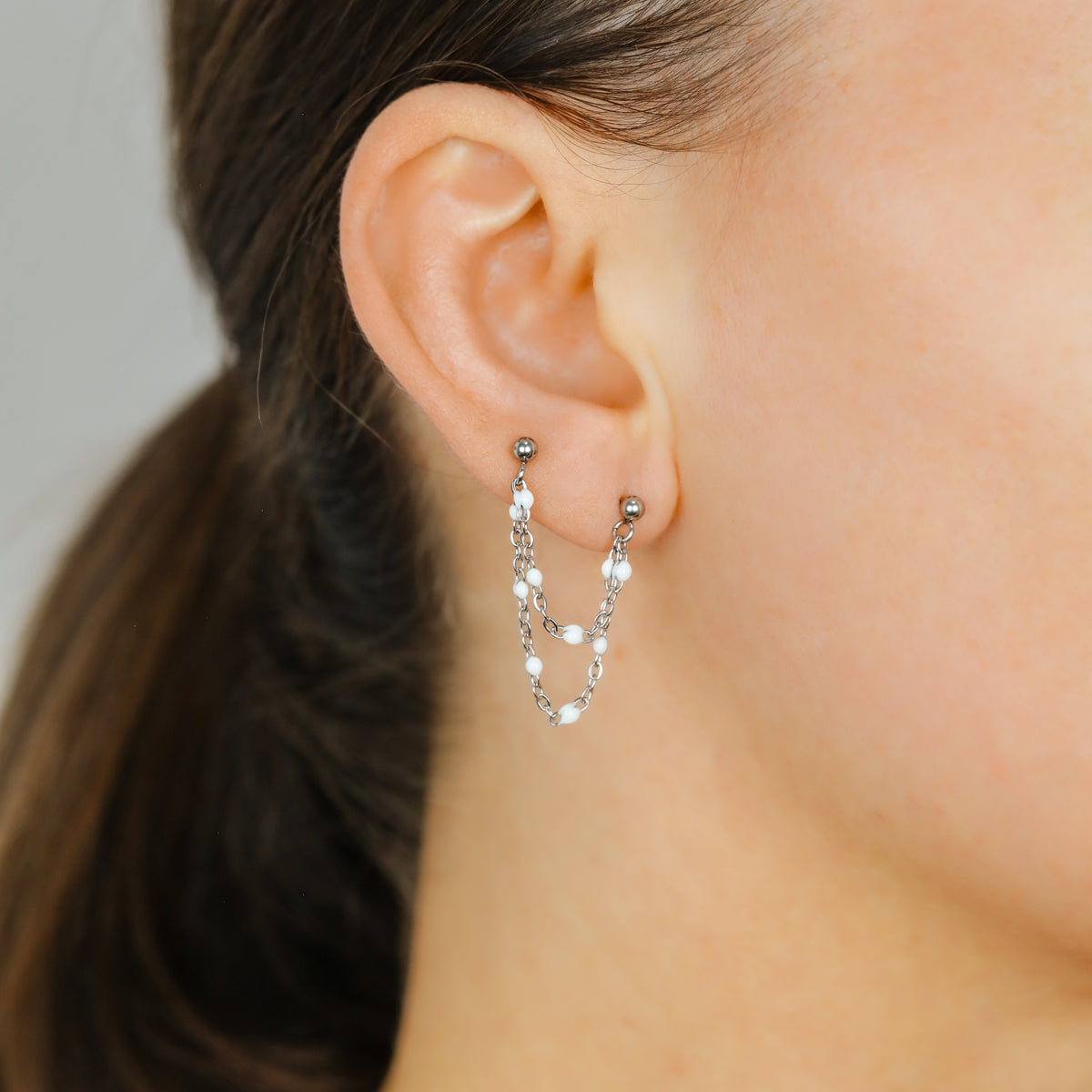 Silver double chain stud earring worn on two lobe piercings with white enamel beads