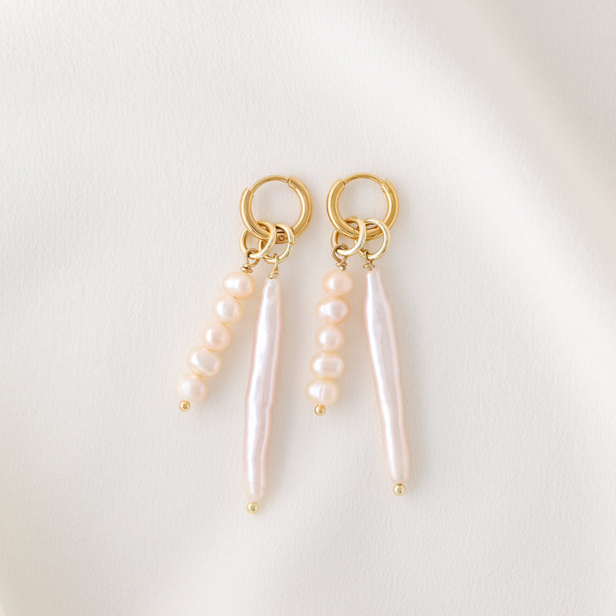 Two gold plated huggie hoop earrings with asymmetrical pink baroque pearl charms resting elegantly on a soft cream-colored silk fabric.