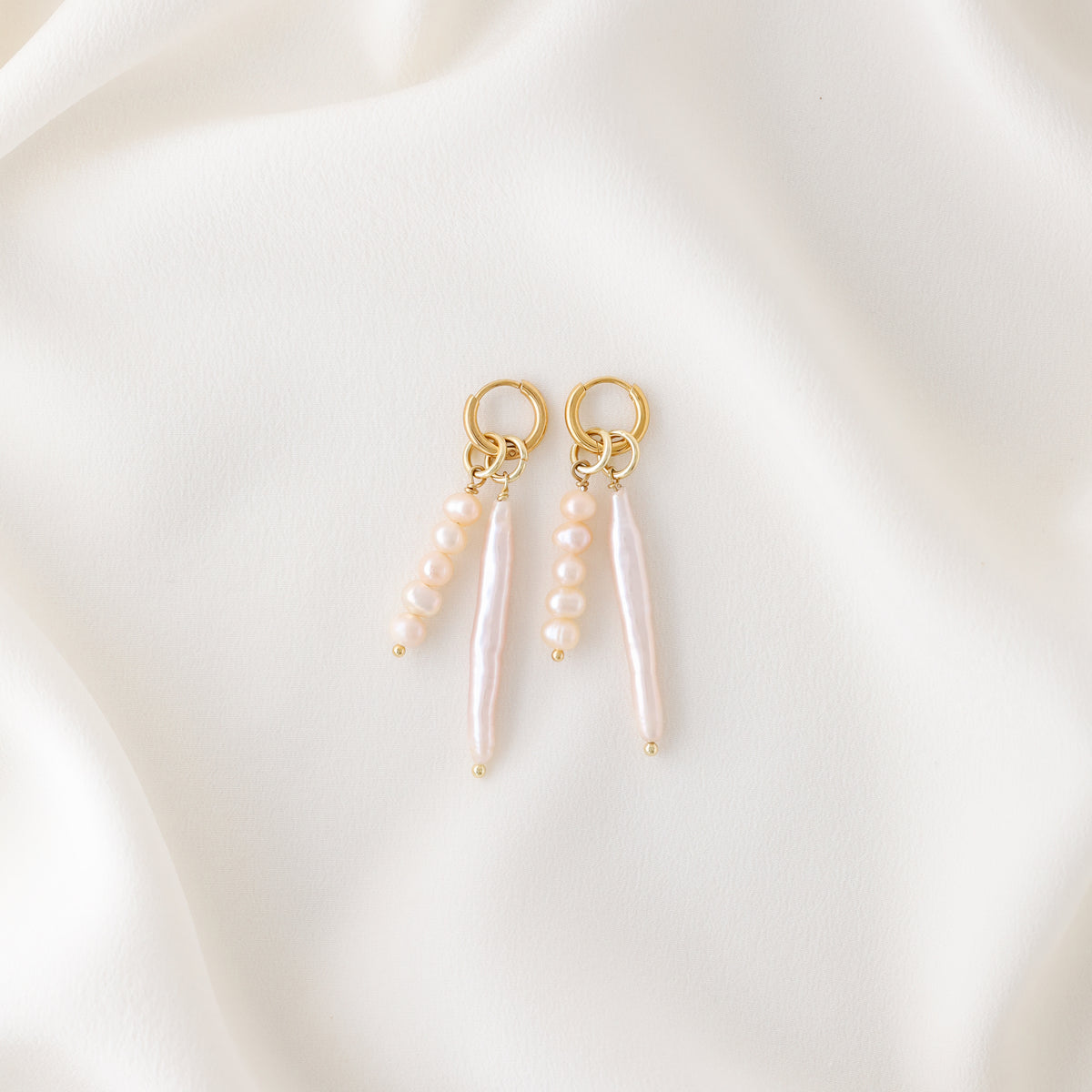 Two gold plated huggie hoop earrings with asymmetrical pink baroque pearl charms resting elegantly on a soft cream-colored silk fabric.