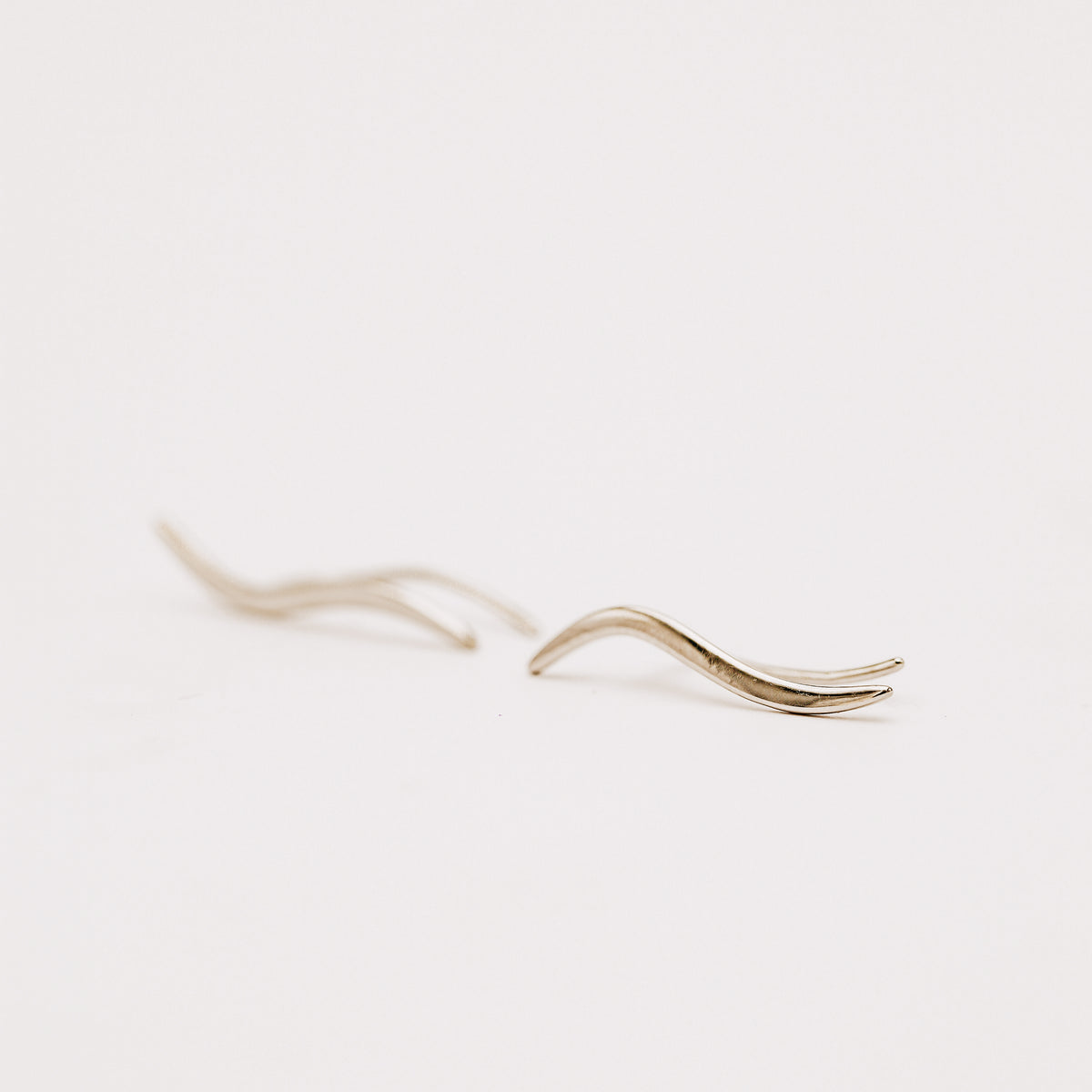 Matching left and right rhodium plated wave ear climbers displayed on a neutral stone surface, showing the mirrored curved silhouettes and high-polish finish.