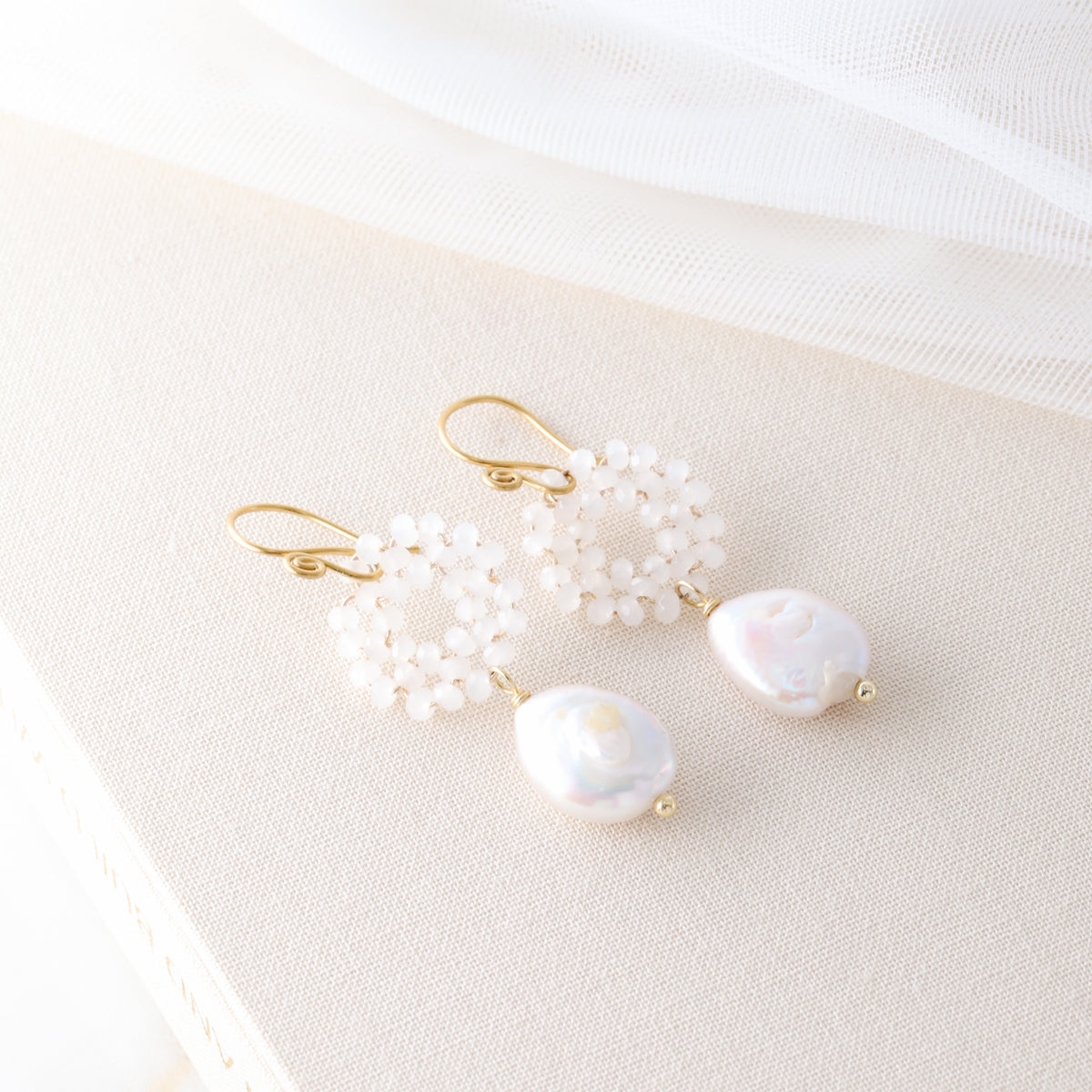 A pair of gold-plated baroque freshwater pearl earrings with woven glass crystal wreaths resting on a textured cream-colored book.