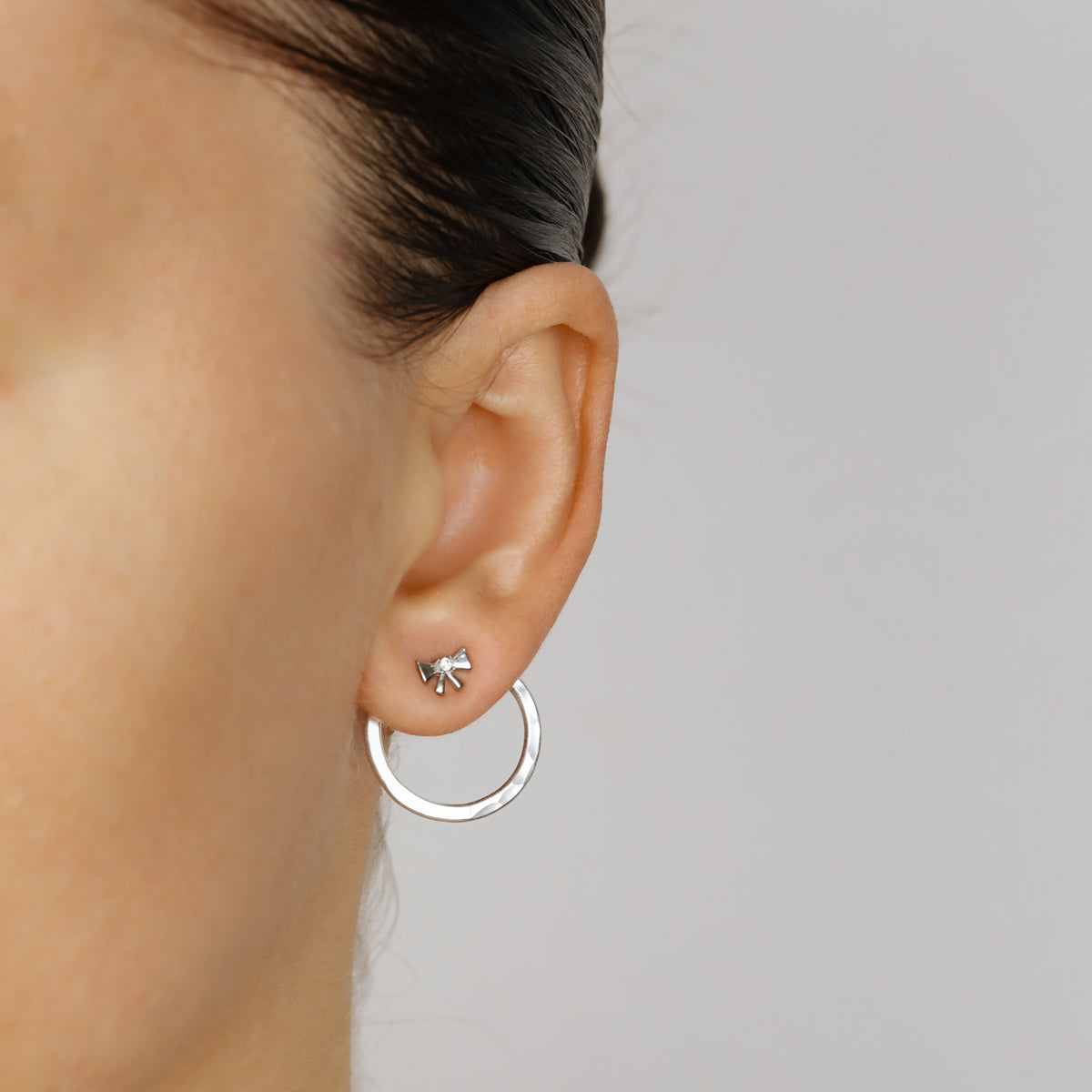 Silver round ear jacket worn behind the lobe with a small bow stud, showing the circular hammered jacket hanging below the ear.