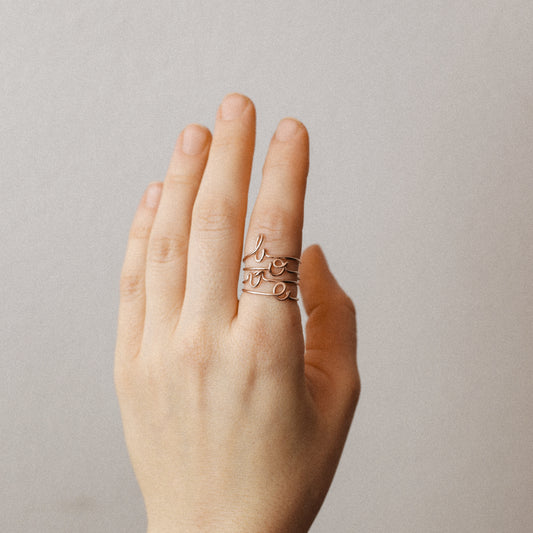 Close-up of a handmade 14k rose gold filled wire ring shaped into the letter L, O, V and E on a woman's finger.