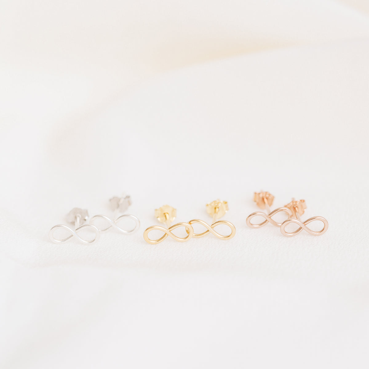 Tiny infinity stud earrings in silver gold and rose gold on soft white fabric