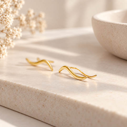 Pair of gold plated wave ear climbers displayed in soft natural light, featuring a flowing curved design with a sleek polished surface.