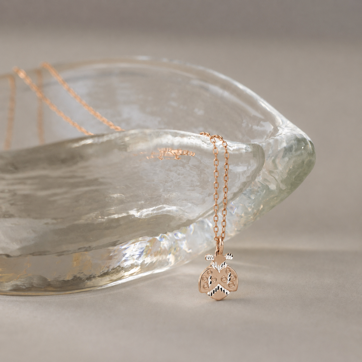 Gold ladybug necklace displayed beside textured glass bowl with soft neutral background