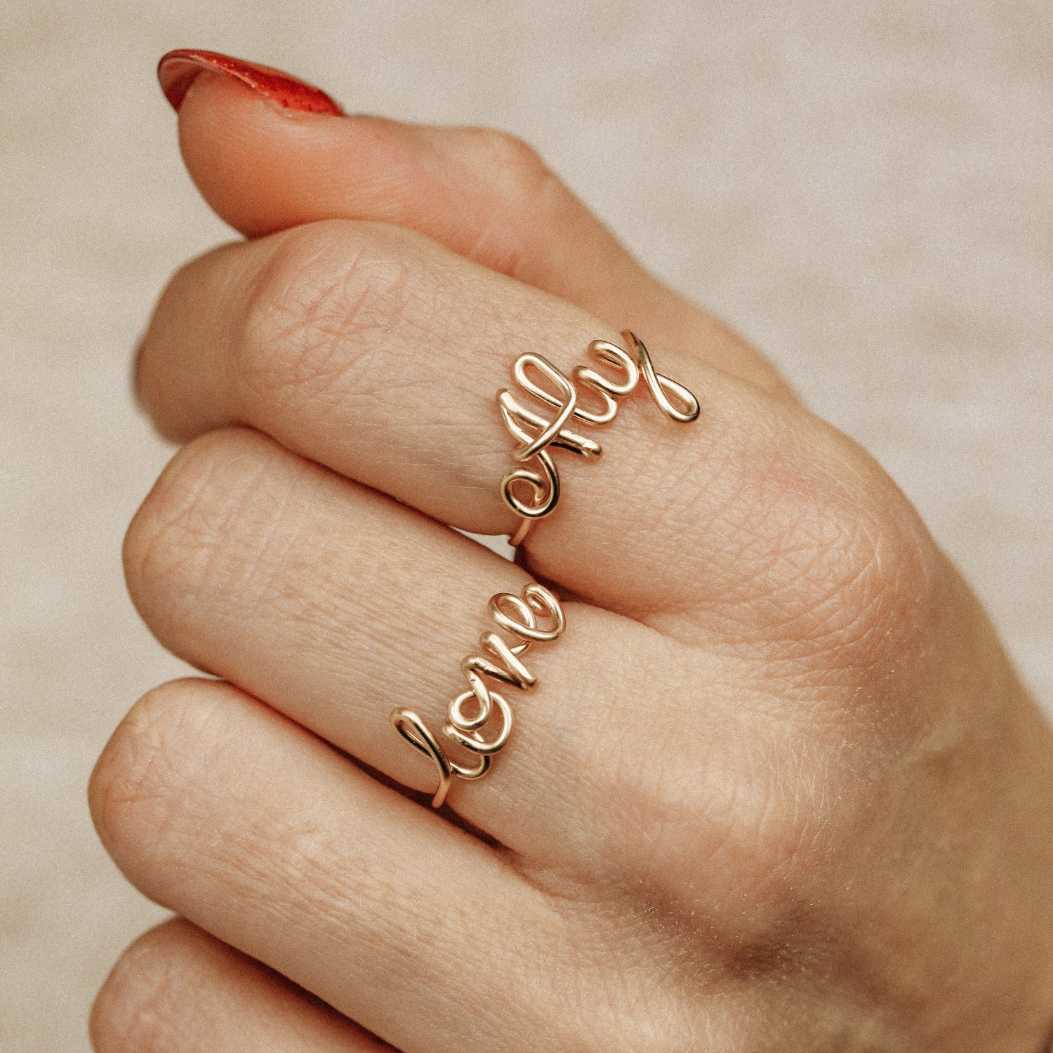 Two handmade gold wire rings featuring the words "Aly" and "Love" in elegant cursive script, worn together on a hand with a red manicure.