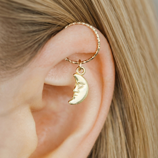 Close-up of a 18k yellow gold vermeil man in the moon charm hanging from a diamond-cut textured gold hoop earring worn in a helix piercing.