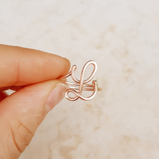 Dainty handmade rose gold wire ring featuring an uppercase cursive letter L held between a thumb and forefinger.