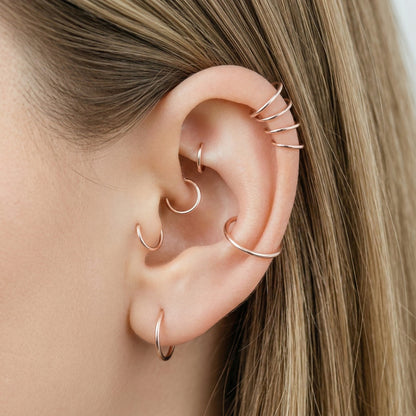 Close-up of a human ear featuring multiple smooth rose gold seamless hoop earrings in various sizes, including helix, tragus, and lobe piercings.