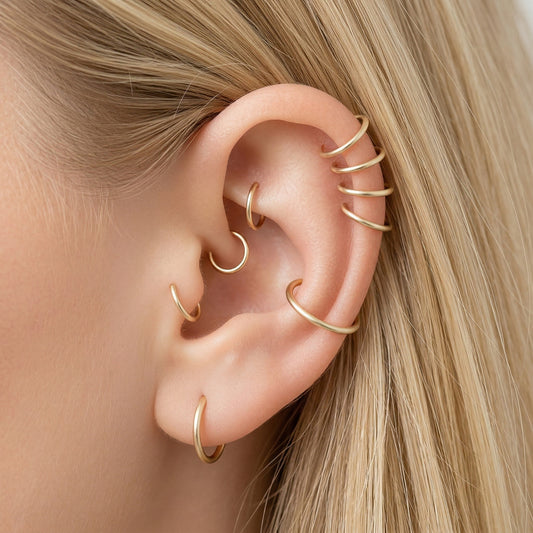 Close-up of a human ear featuring multiple smooth gold seamless hoop earrings in helix, tragus, and lobe piercings against a neutral background.