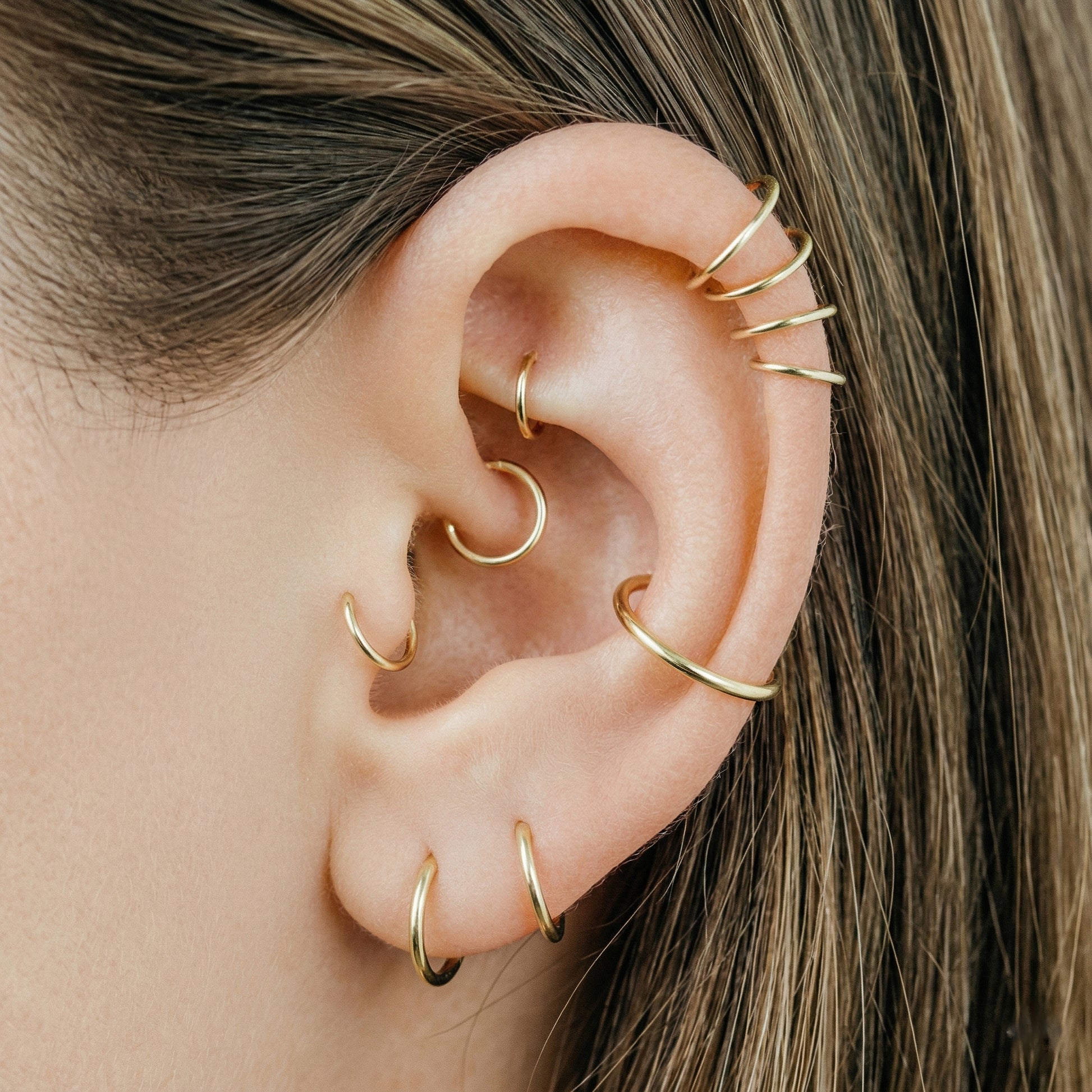 Close-up of a human ear featuring multiple smooth gold seamless hoop earrings in helix, tragus, and lobe piercings against a neutral background.