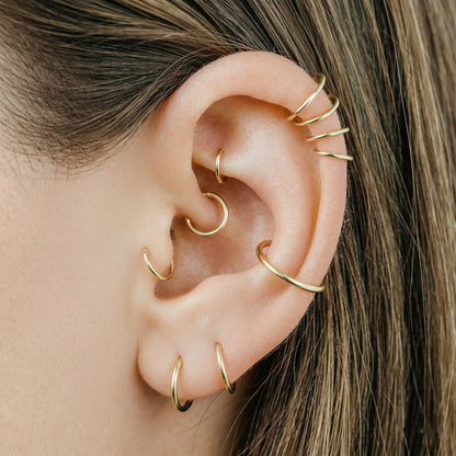 Close-up of a human ear featuring multiple smooth gold seamless hoop earrings in helix, tragus, and lobe piercings against a neutral background.