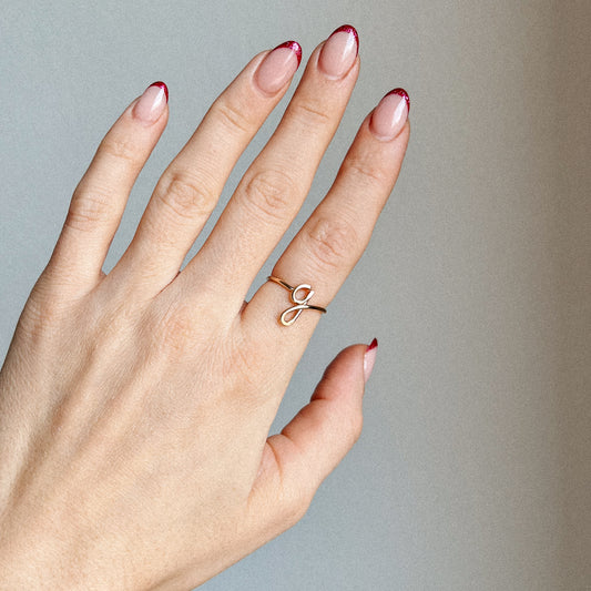 Close-up of a handmade 14k gold filled wire ring shaped into the letter G on a woman's finger with red French tip nails.