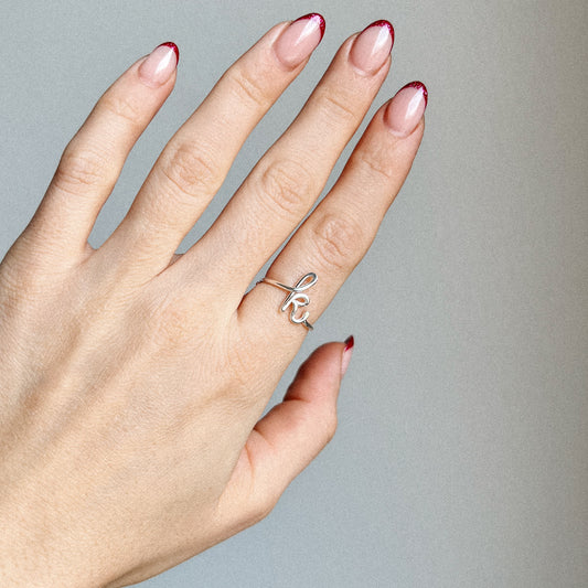 Close-up of a delicate handmade gold wire ring shaped into a cursive lowercase letter k on a woman's finger with red French tip nails.
