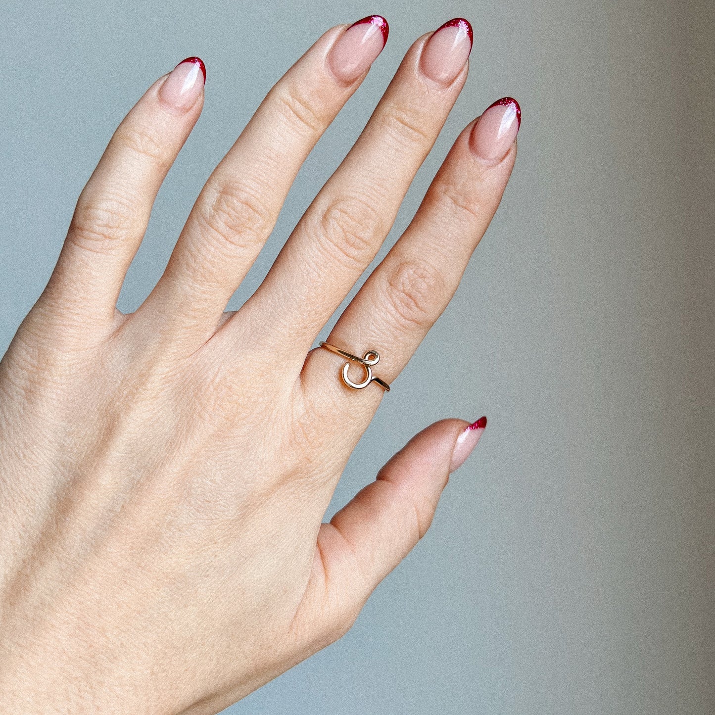 Close-up of a handmade 14k gold filled wire ring shaped into the letter S on a woman's finger with red French tip nails.