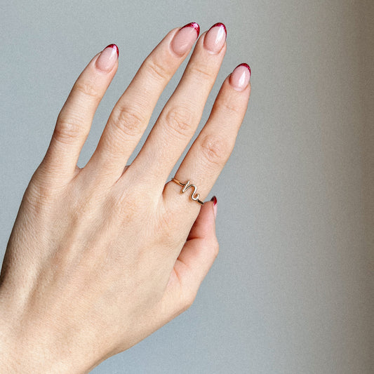 Close-up of a handmade 14k gold filled wire ring shaped into the letter N on a woman's finger with red French tip nails.