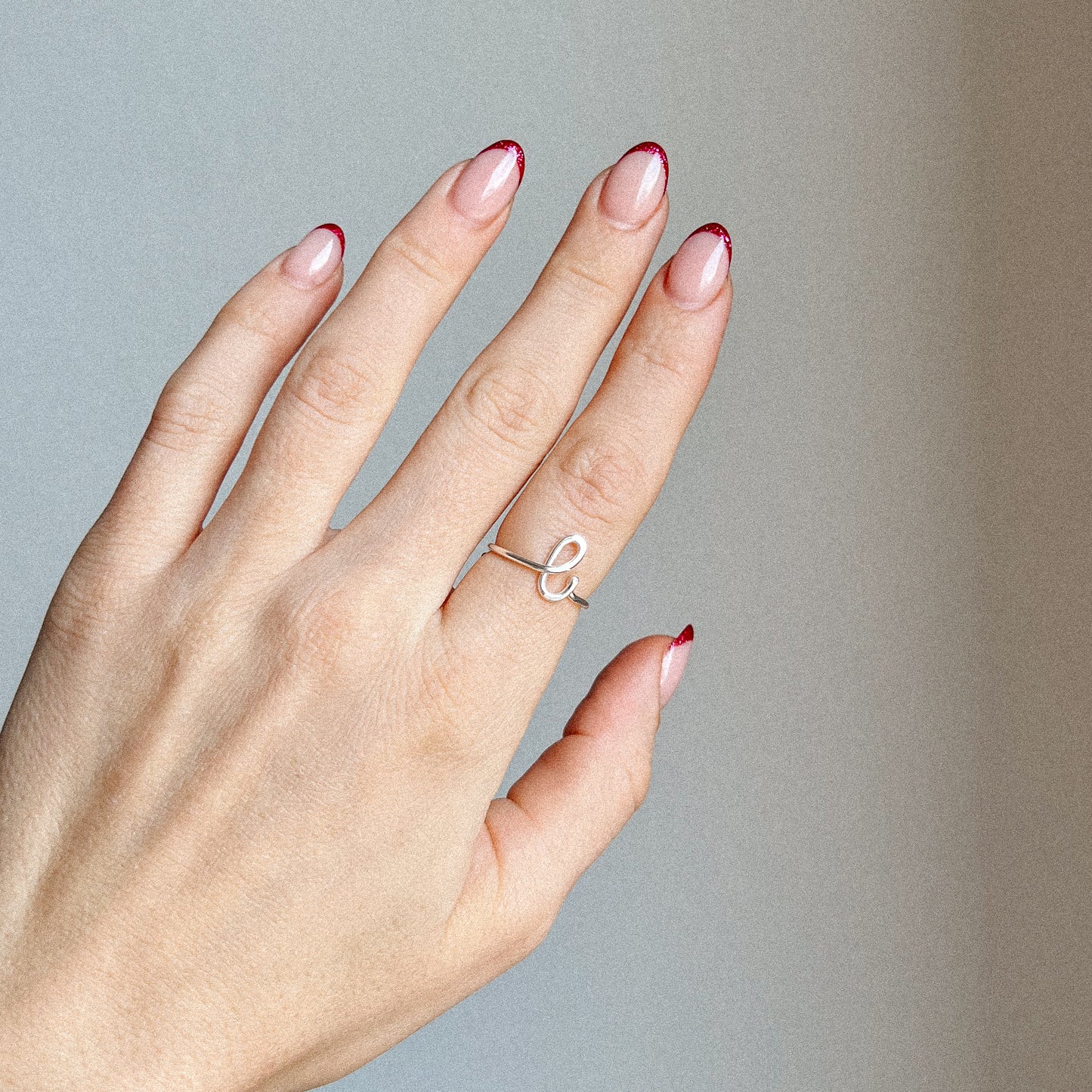 Close-up of a handmade 925 sterling silver wire ring shaped into the letter L on a woman's finger with red French tip nails.