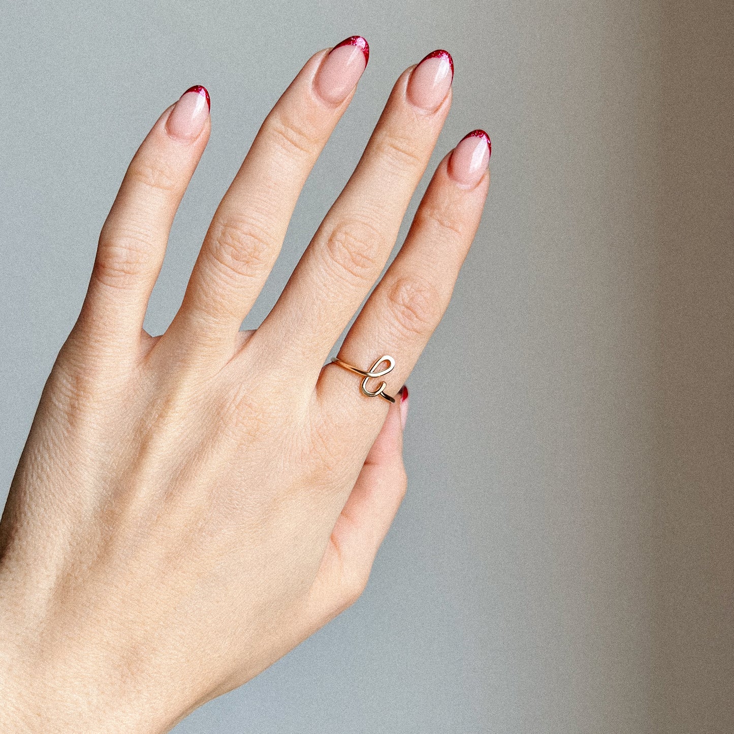 Close-up of a handmade 14k gold filled wire ring shaped into the letter L on a woman's finger with red French tip nails.