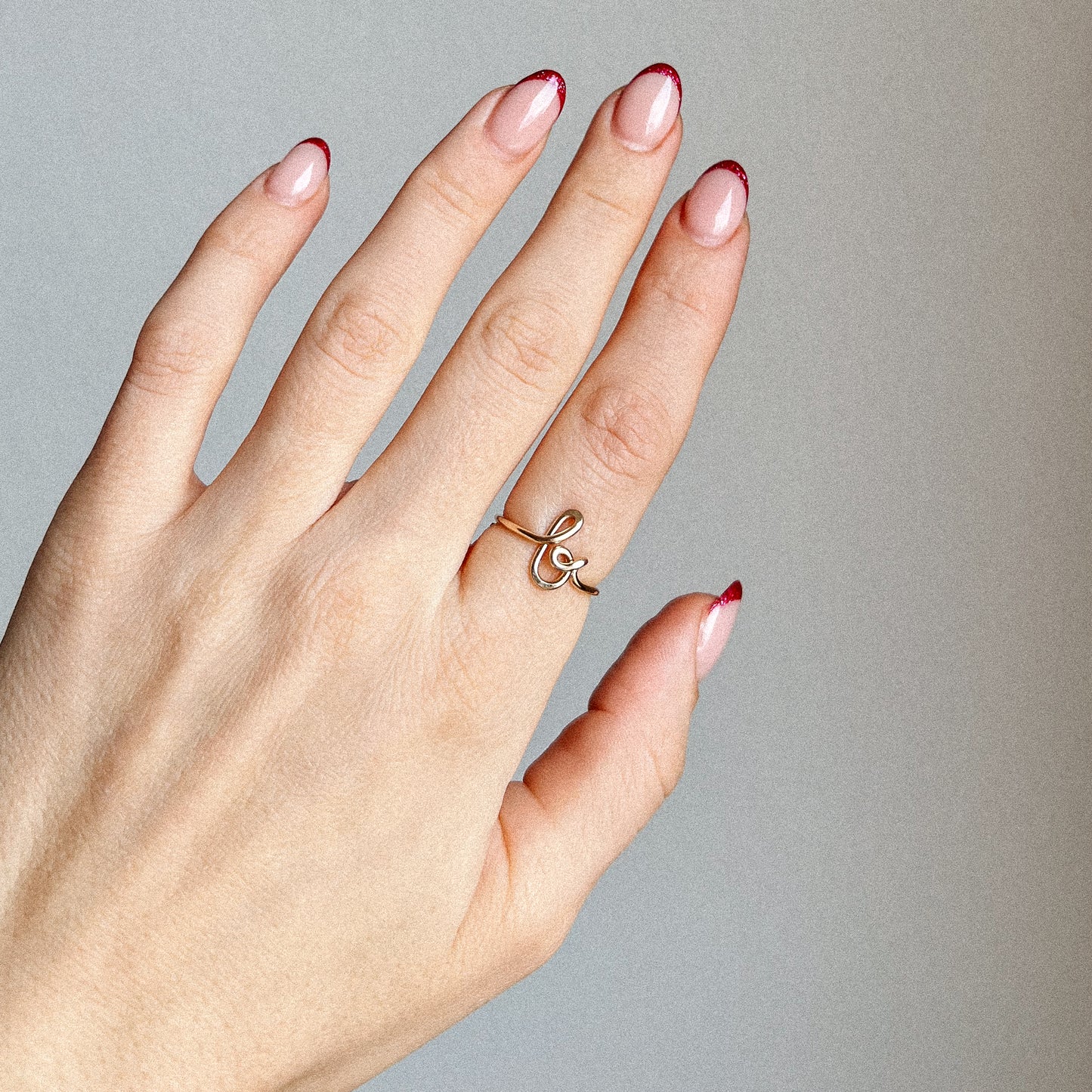 Close-up of a handmade 14k gold filled wire ring shaped into the letter B on a woman's finger with red French tip nails.