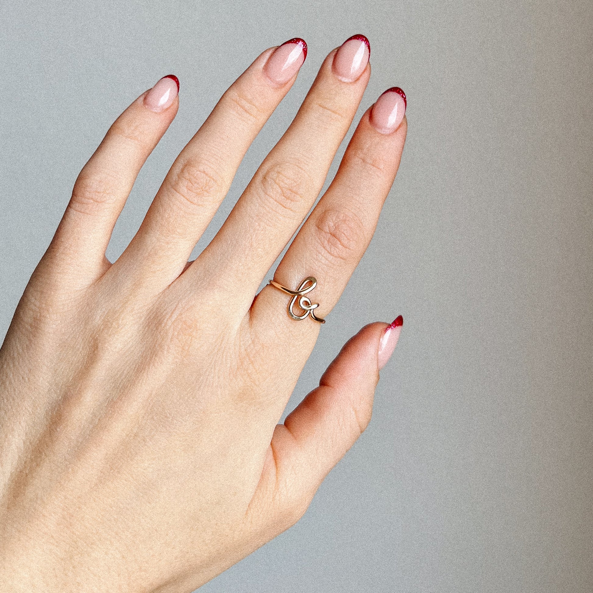 Close-up of a handmade 14k gold filled wire ring shaped into the letter B on a woman's finger with red French tip nails.