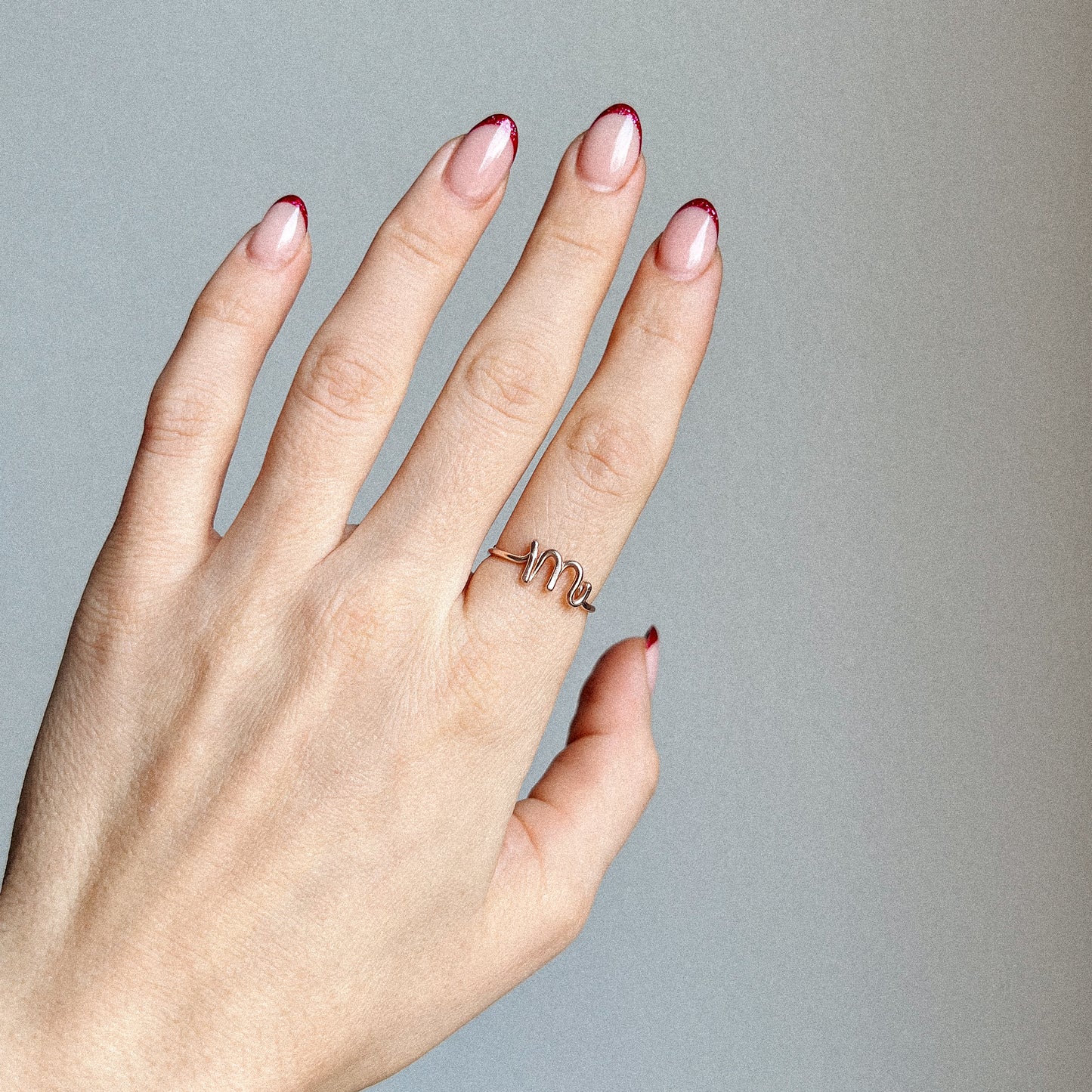 Close-up of a handmade 14k gold filled wire ring shaped into the letter M on a woman's finger with red French tip nails.