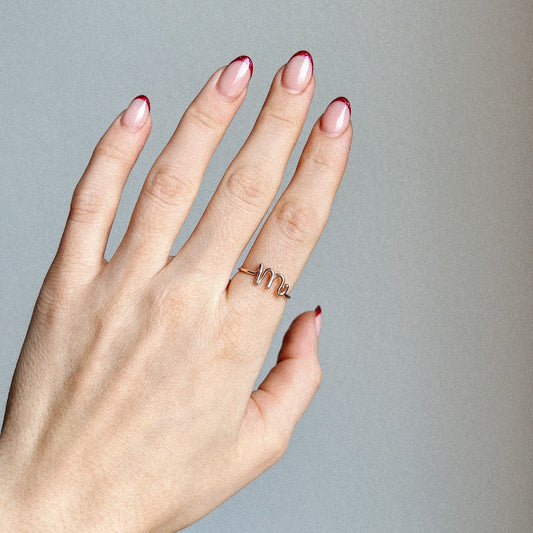 Close-up of a handmade 14k gold filled wire ring shaped into the letter M on a woman's finger with red French tip nails.