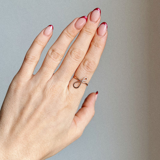 Handcrafted adjustable gold ring featuring a dainty cursive lowercase letter F on a woman's hand with red tipped french manicure.