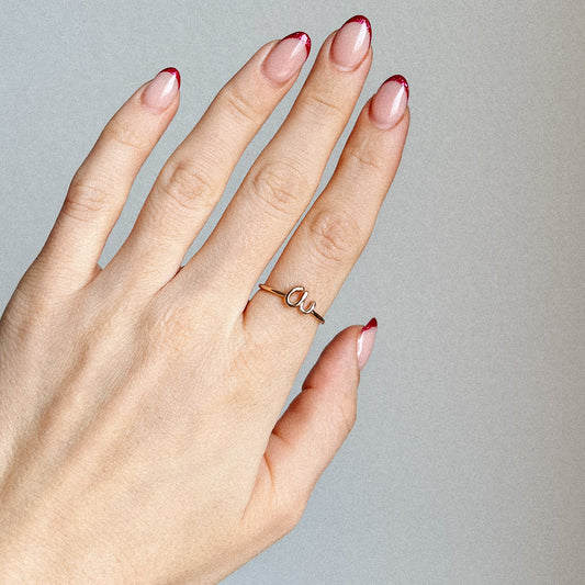 Hand holding a delicate gold wire ring featuring a lowercase cursive letter 'a' with a minimalist adjustable band and red French tip nails.