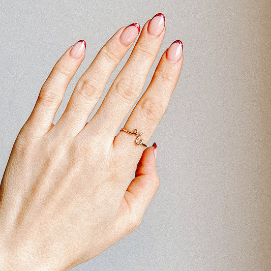 Close-up of a handmade 14k gold filled wire ring shaped into the letter R on a woman's finger with red French tip nails.