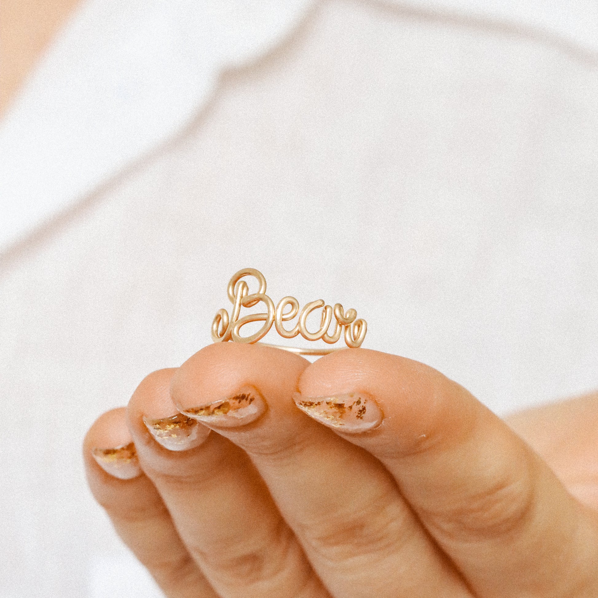 Custom gold wire ring with the name "Bear" in elegant script, styled on a hand with a white shirt.