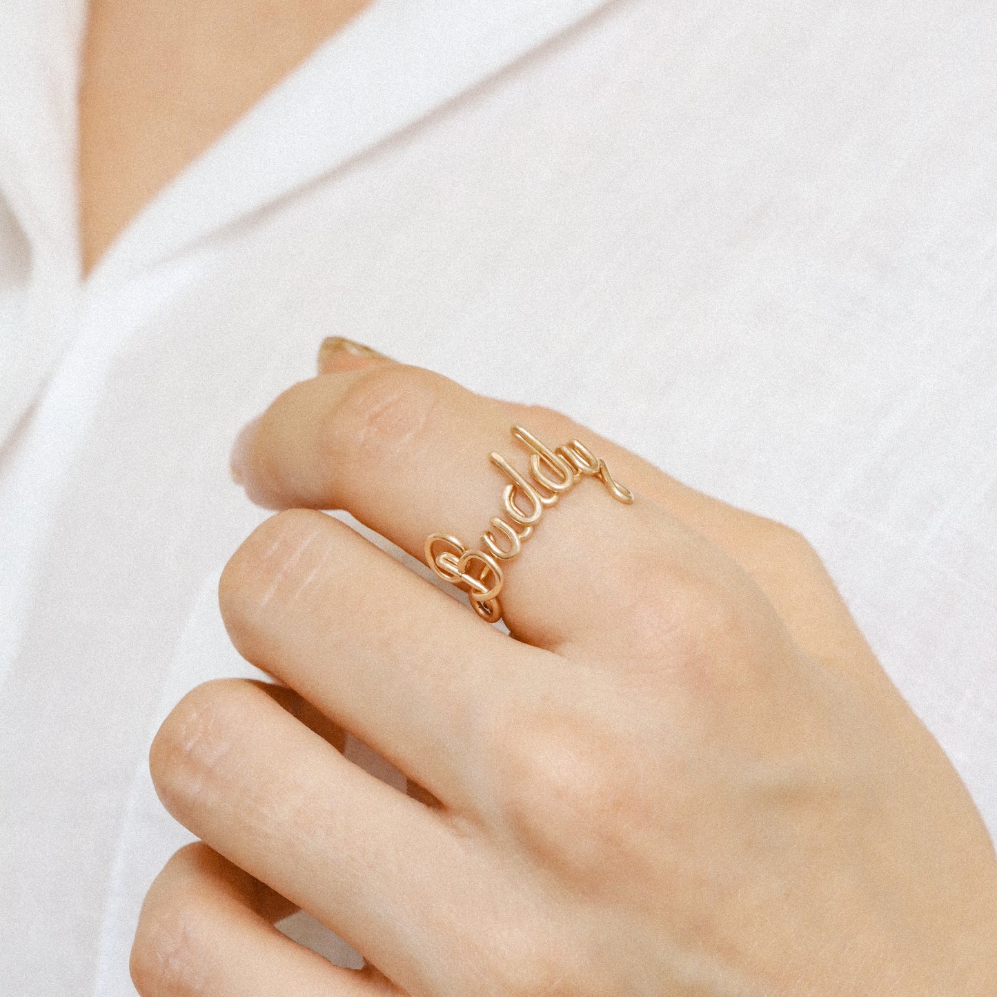 Custom gold wire ring with the name "Buddy" in elegant script, styled on a hand with a white shirt.