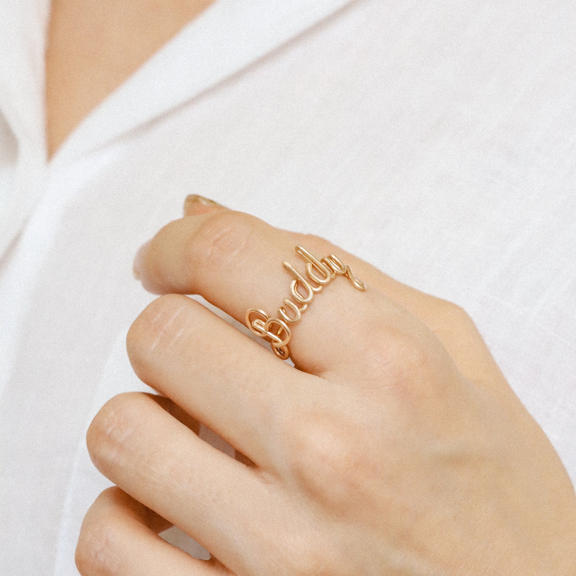 Custom gold wire ring with the name "Buddy" in elegant script, styled on a hand with a white shirt.
