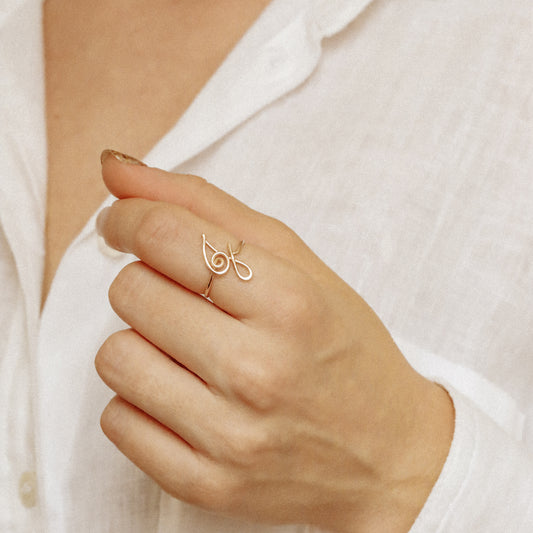 Handmade gold wire ring with an uppercase cursive letter K, worn on a hand against a white linen shirt.