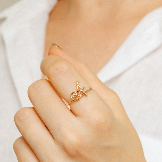 Dainty handmade gold wire ring with an uppercase cursive letter A, worn on a hand against a white shirt.