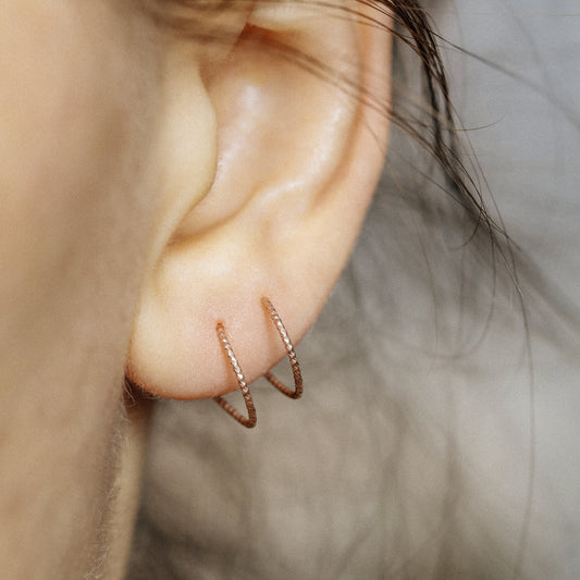 Close-up of a woman's ear wearing tiny rose gold double hoop illusion earrings with a diamond-cut texture, designed to create the appearance of two piercings with a single hole.