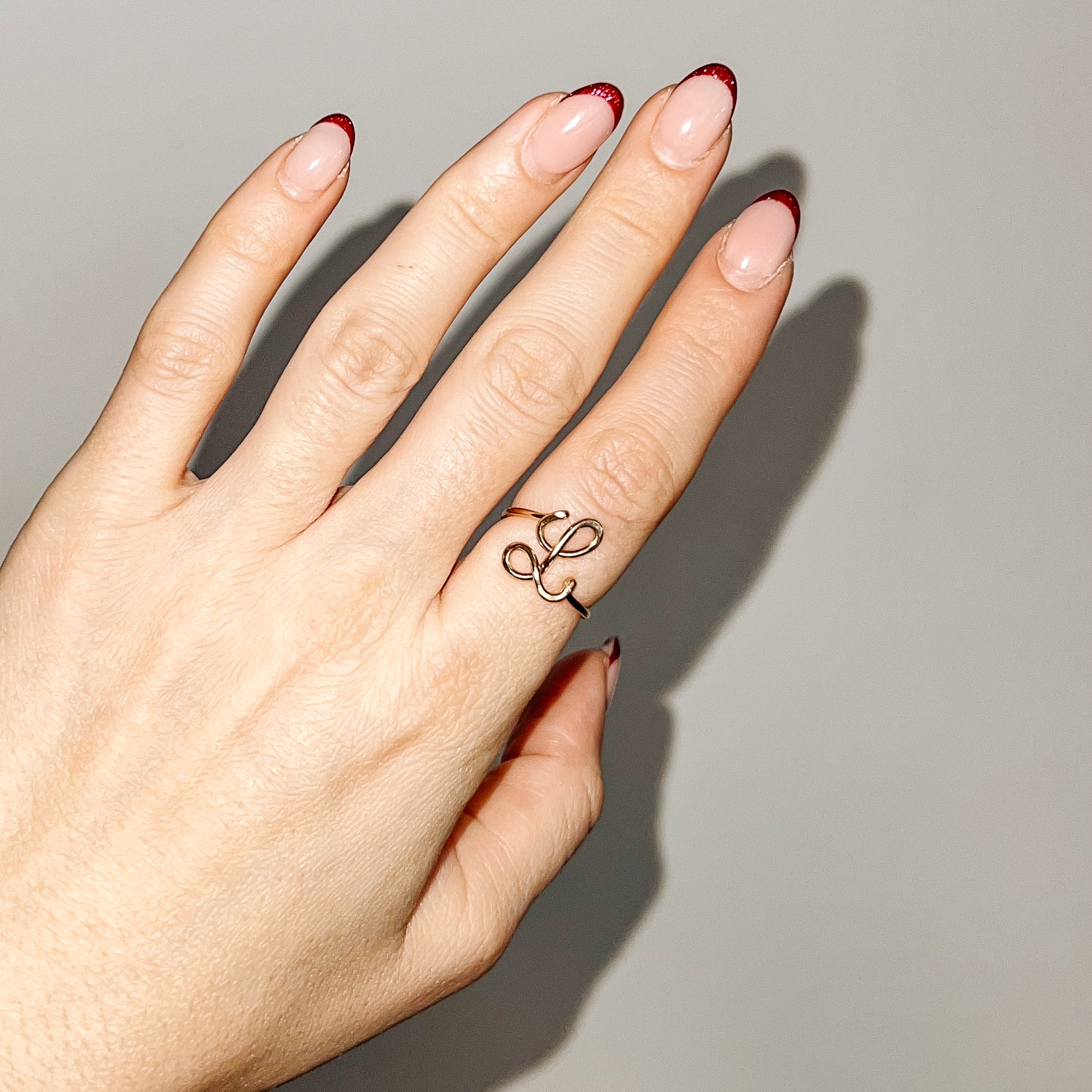Handmade gold wire ring with an uppercase cursive letter L design, styled on a hand with red French manicure.