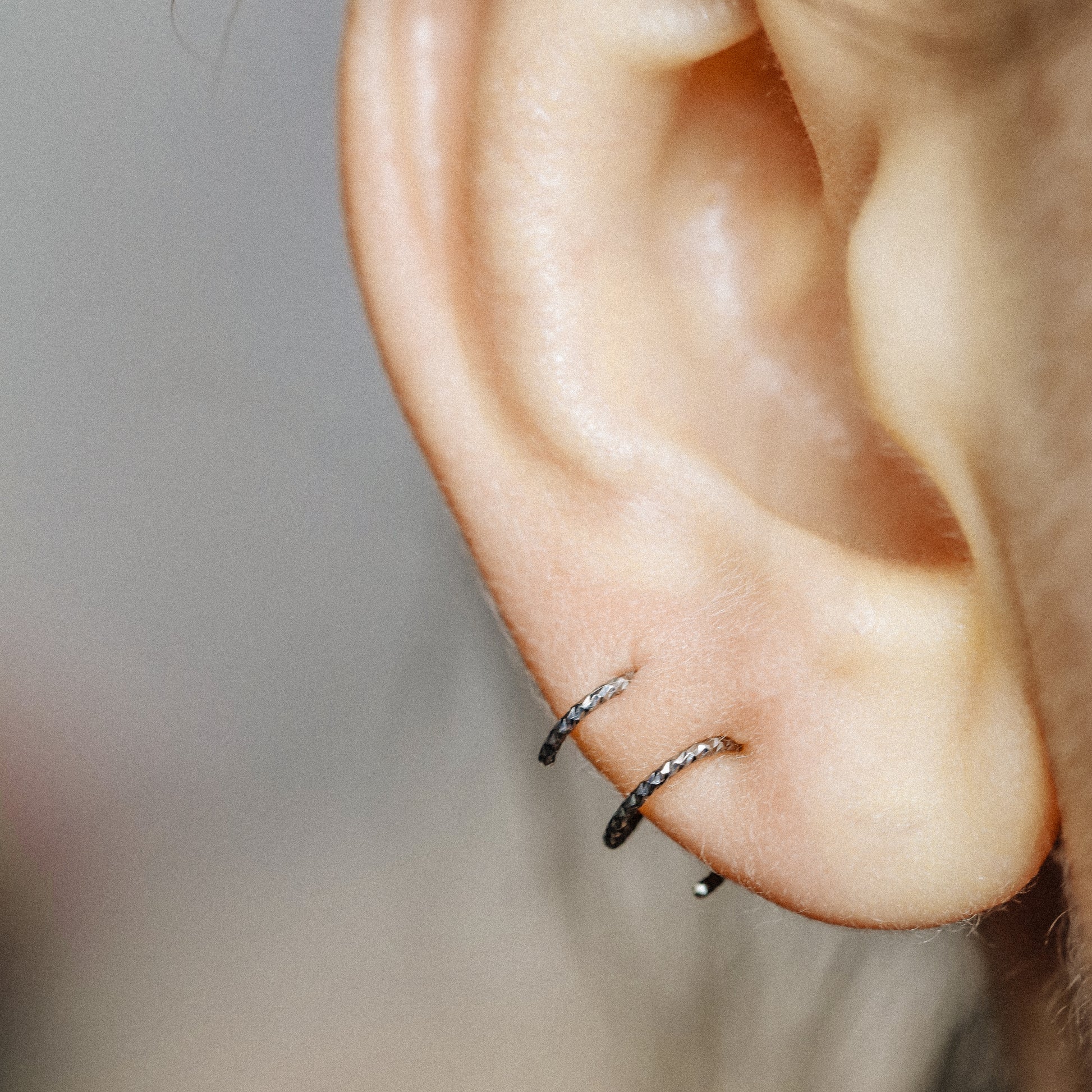 Minimal black double hoop spiral earrings worn on ear, tiny ruthenium sterling silver illusion hoops creating double piercing look, diamond cut texture close-up