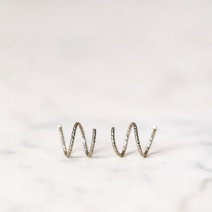 Minimal black double hoop spiral earrings worn on ear, tiny ruthenium sterling silver illusion hoops creating double piercing look, diamond cut texture close-up